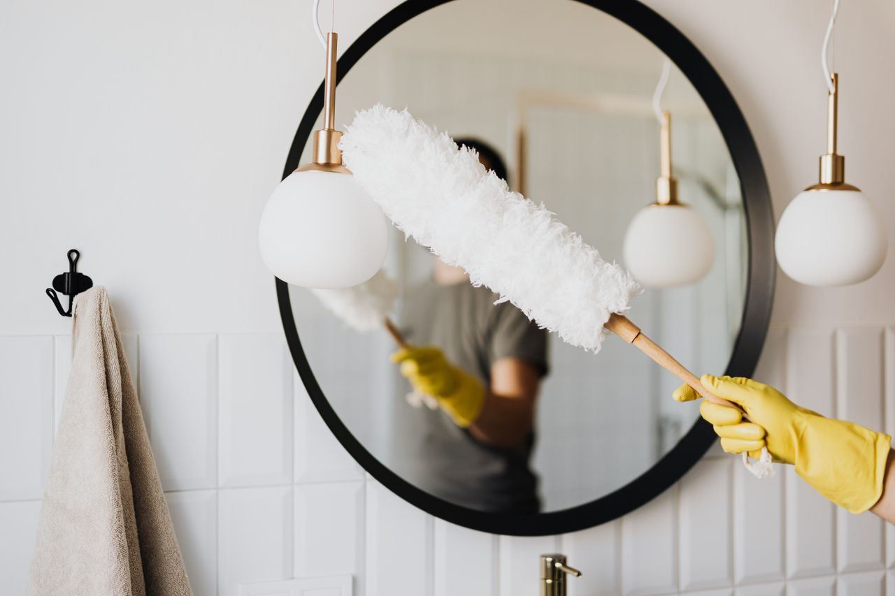 creative-approach-img Donna anonima ritagliata, casalinga con guanti di gomma che spolvera una lampada moderna con una spazzola morbida davanti a uno specchio mentre pulisce un bagno elegante